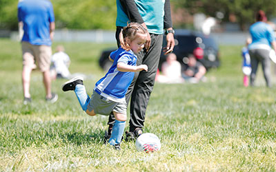 A young girl kicking a soccer ball with YMCA volunteer nearby
